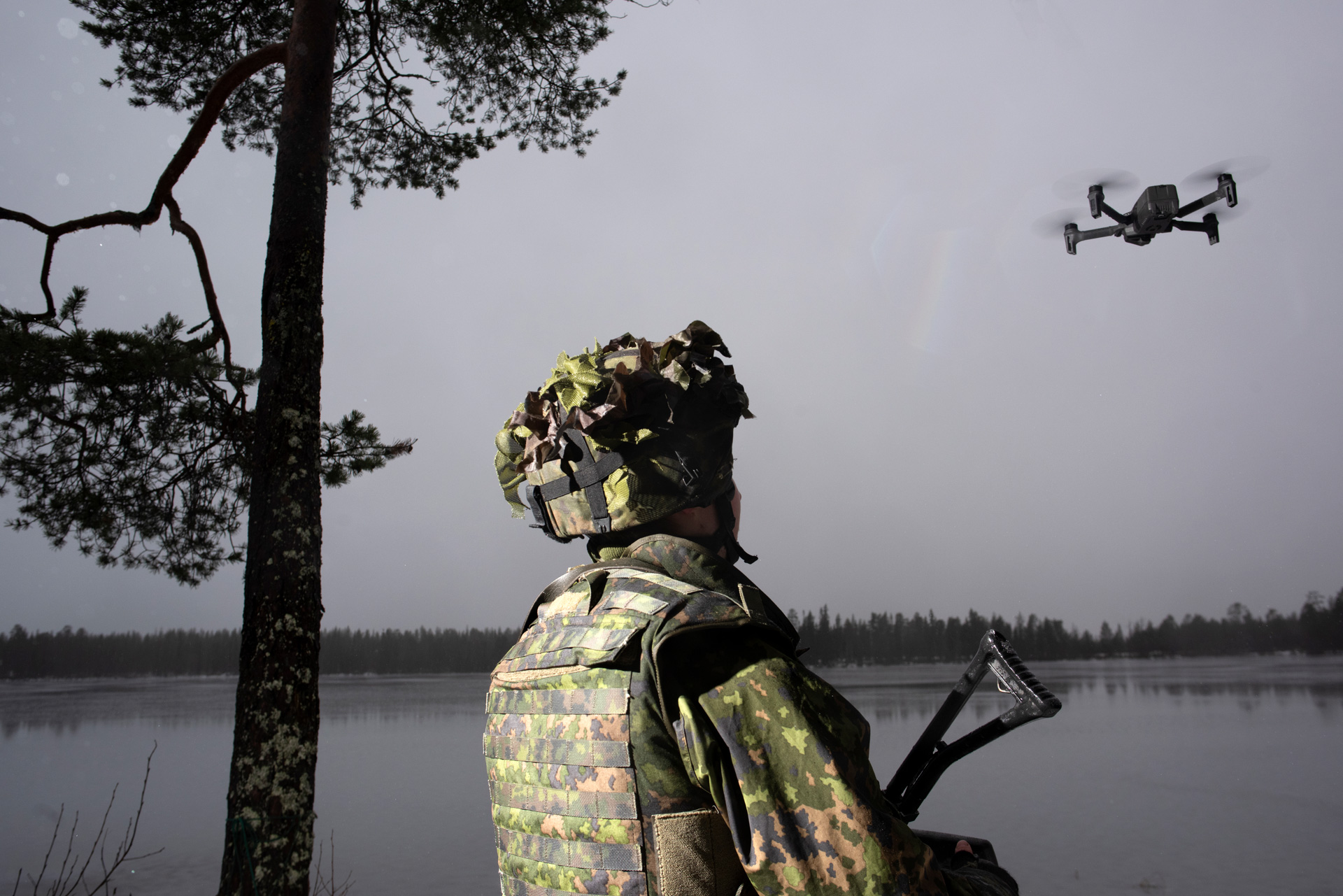 The upper half of a Finnish soldier wearing a green and khaki camouflage uniform and helmet is framed, with their face facing up and away from the camera. They are looking up at a small quad-copter drone hovering just above them. The stock of the soldier's weapon can be glimpsed to one side. On the other stands a tall tree, all against the backdrop of a calm, gray lake ringed by trees and an overcast sky.