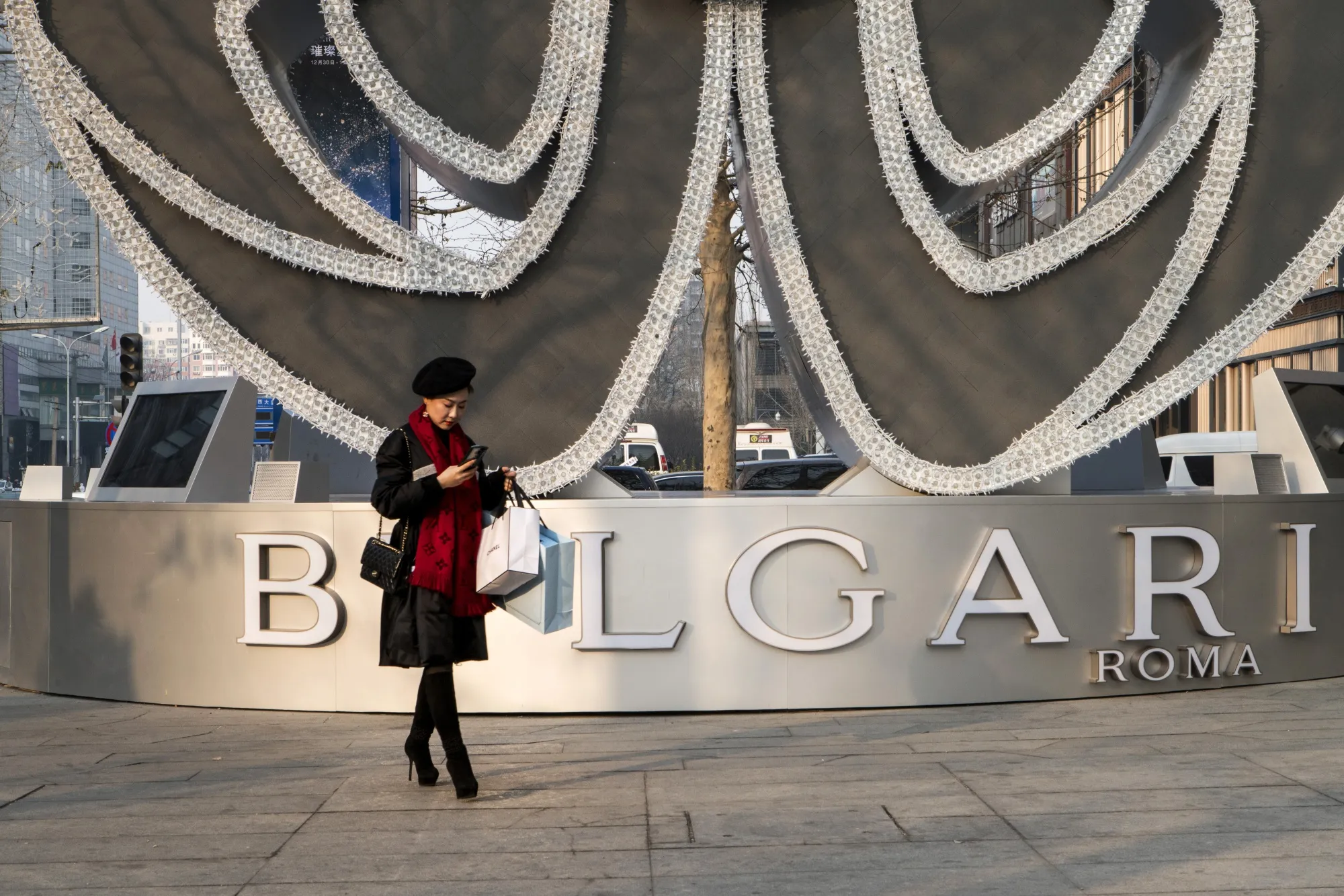 A shopper carrying bags uses a smartphone in front of a Bulgari SpA installation in the Dawanglu area of Beijing.