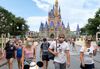 Photograph of six people walking in Walt Disney World, wearing face masks, dressed in T-shirts and shorts. More people walking, sitting or taking photographs can be seen in the background, along with Disney’s flagship Cinderella Castle, a large castle with blue-purple spires and blush-colored towers.