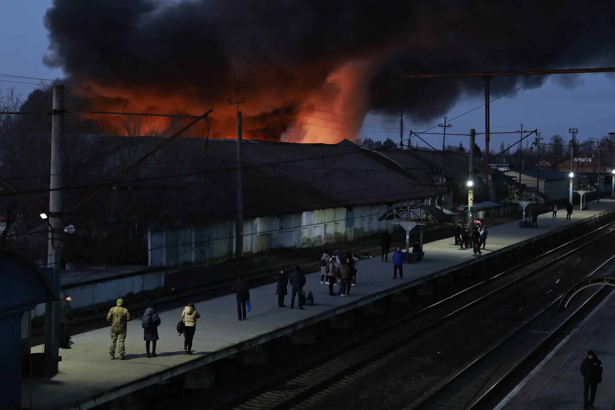 People wait for a train on a platform in front of a fire in a residential area caused by a Russian missile strike&nbsp;in Brovary, Ukraine on March 14.&nbsp;