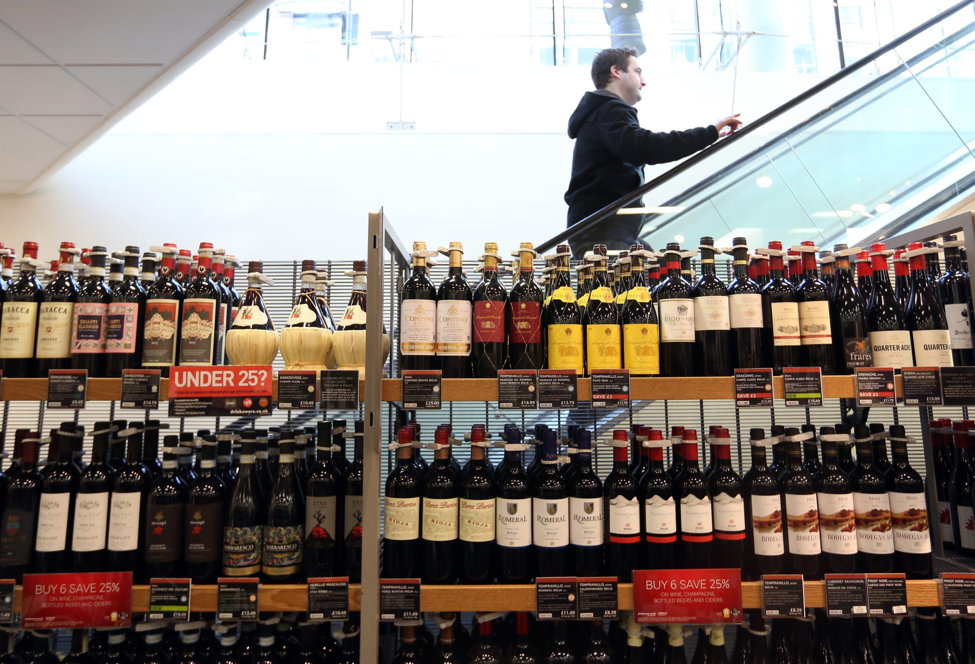 A customer ascends an escalator behind a shelf displaying bottles of wine in a supermarket.&nbsp;