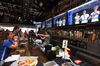 The inside of a bar, where soccer fans are looking at TV screens. A bartender is operating the till behind the bar.