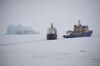 A picture of an icebreaker opening a path for a cargo ship. There is an iceberg in the background. The landscape is all snow and ice, and the sky is foggy.