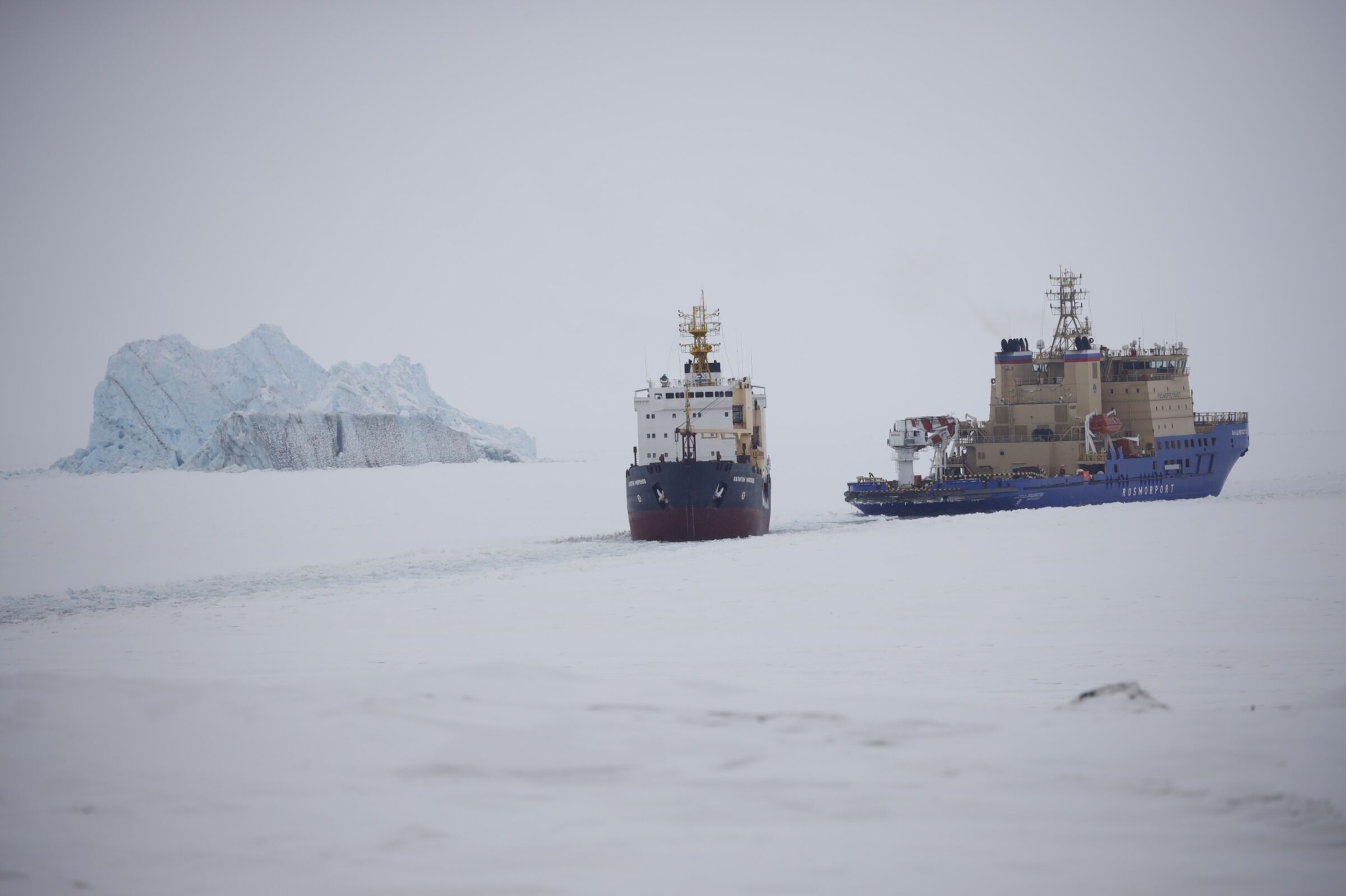Egy jégtörő képe, amely utat nyit egy teherhajónak.  Jéghegy van a háttérben.  A táj csupa hó és jég, az ég ködös.