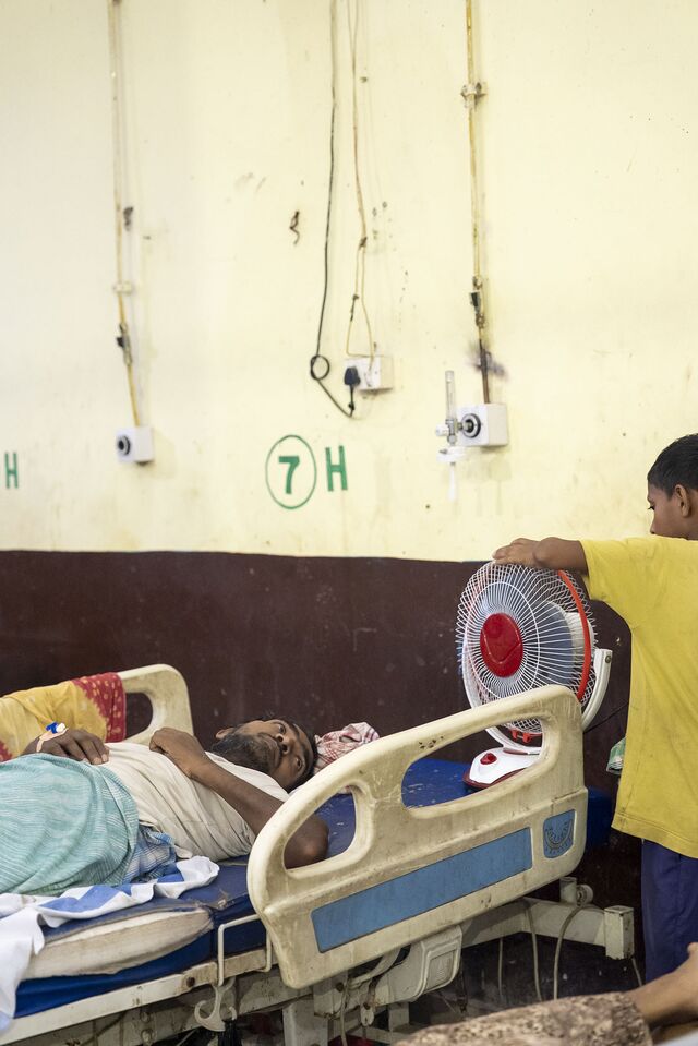 A son adjusts the direction of the fan for his father in the emergency ward at ANMMCH