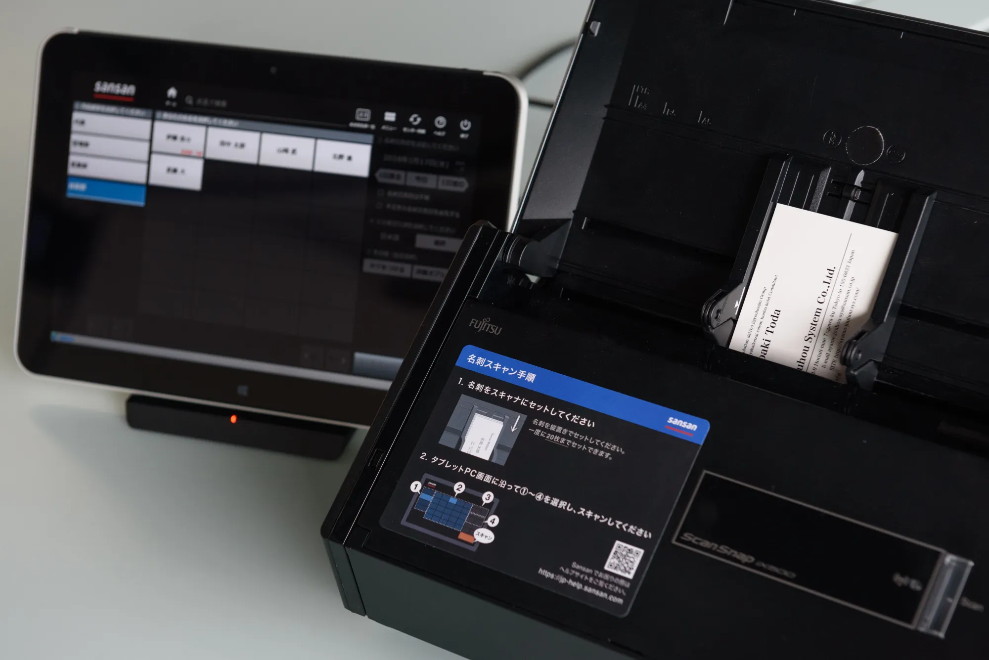 A tablet displaying a sample page of Sansan Inc.'s database, left, and a Fujitsu Ltd. ScanSnap iX500 Sansan Edition business card scanner.