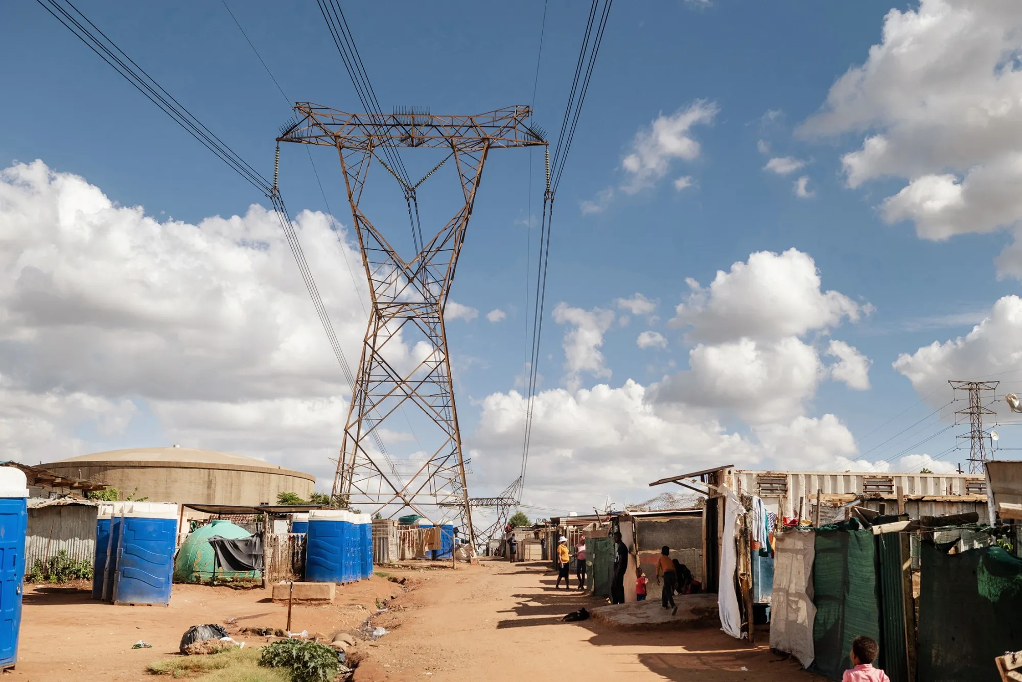 A tall electrical transmission tower rises above homes and portable toilets in the Ivory Park settlement in Johannesburg, illustrating limited access to reliable electricity.