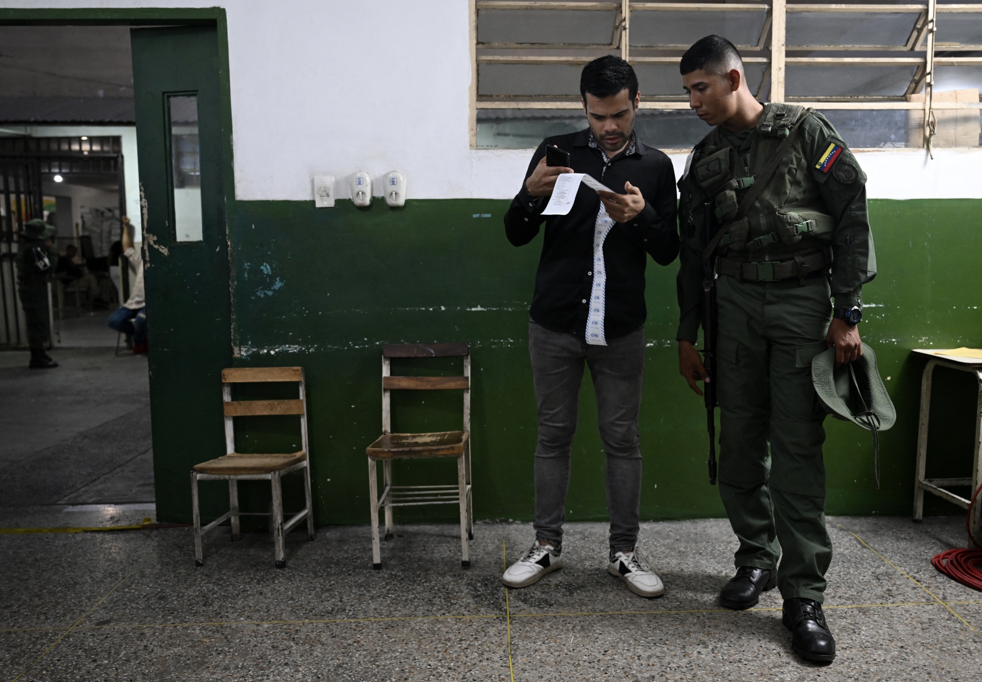 Two men in a room, one casually dressed and the other in a military uniform, standing next to each other and looking at a long thing sheet of paper