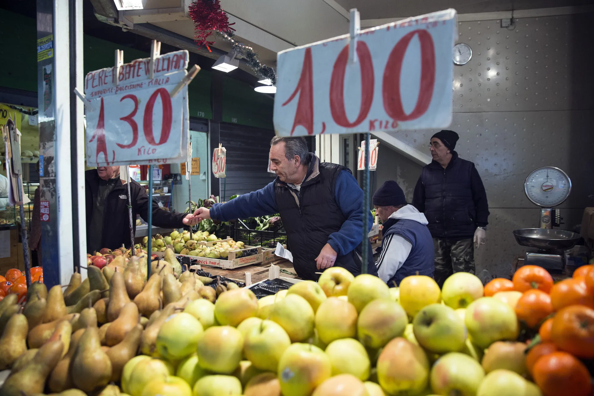 Handwritten euro price signs hang above a display of fruit on a market vendor's stall at an indoor market in Rome.