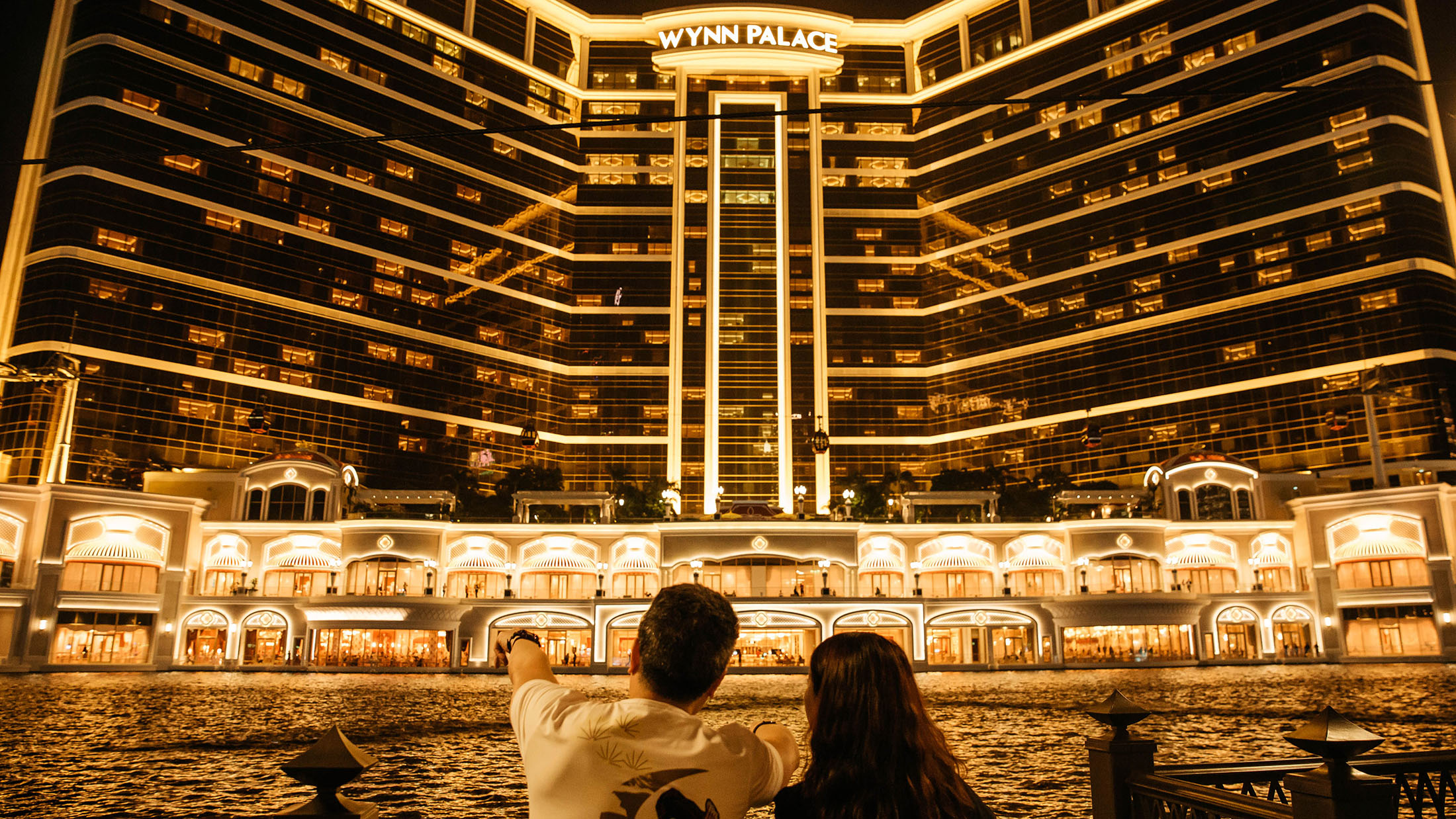 People stand in front of Wynn Resorts Ltd.'s Wynn Palace casino resort at night in Macau, China, on Sunday, Aug. 28, 2016. Macau is scheduled to release gross domestic product (GDP) figures on Aug. 30.

