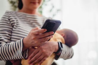 Close up shot of woman carrying her baby and taking a break using smartphone.