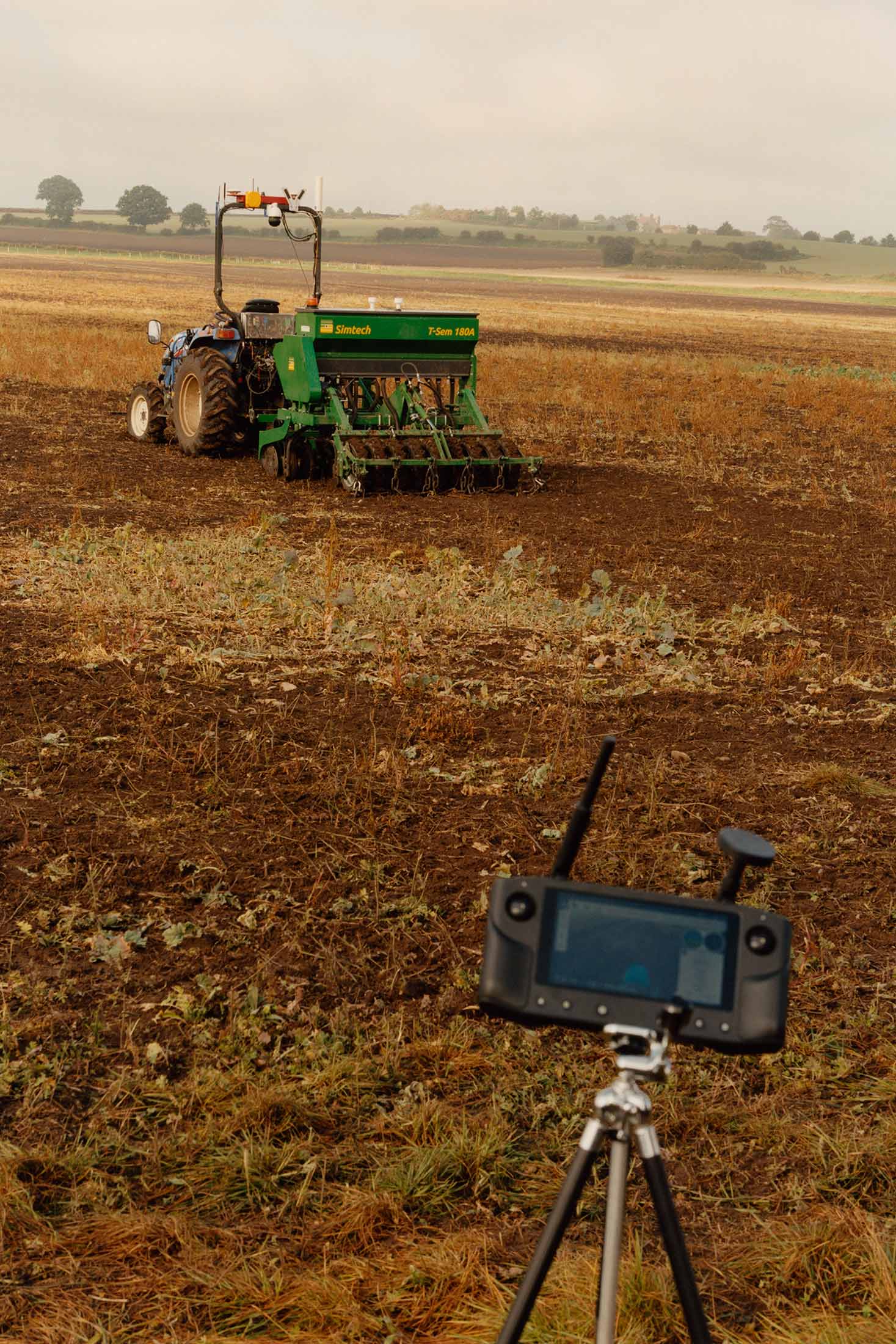 An Iseki tractor tows a seed drill autonomously, with a drone controller in the foreground.