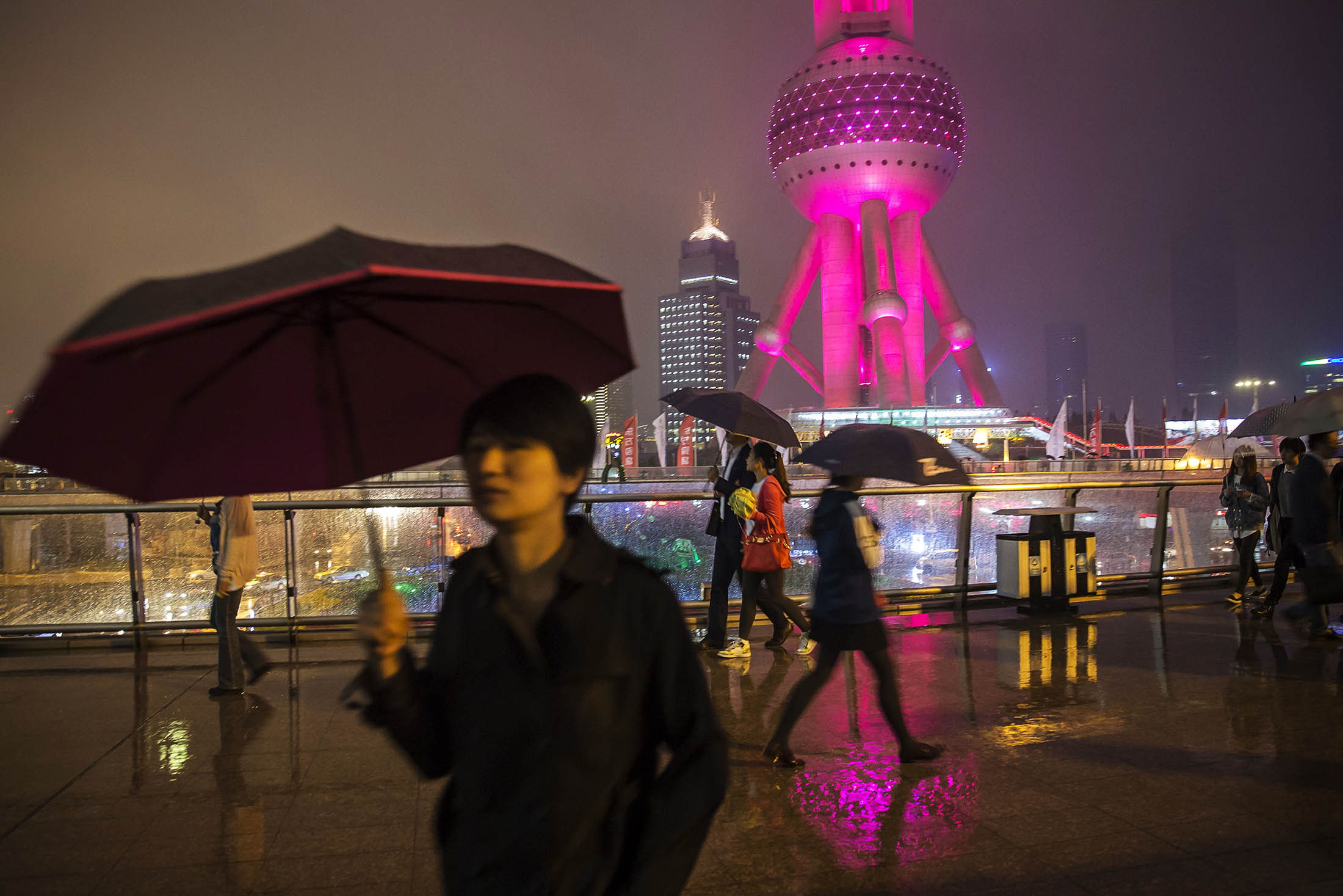 Pedestrians holding umbrellas walk as the Oriental Pearl Tower, right, stands illuminated at night in the Lujiazui district of Shanghai.

