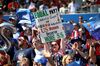 A woman sitting among other soccer fans is holding up a sign reading 'Equal Pay? Three World Cups and Four Gold Medals. US Women Deserve More Dollars'.