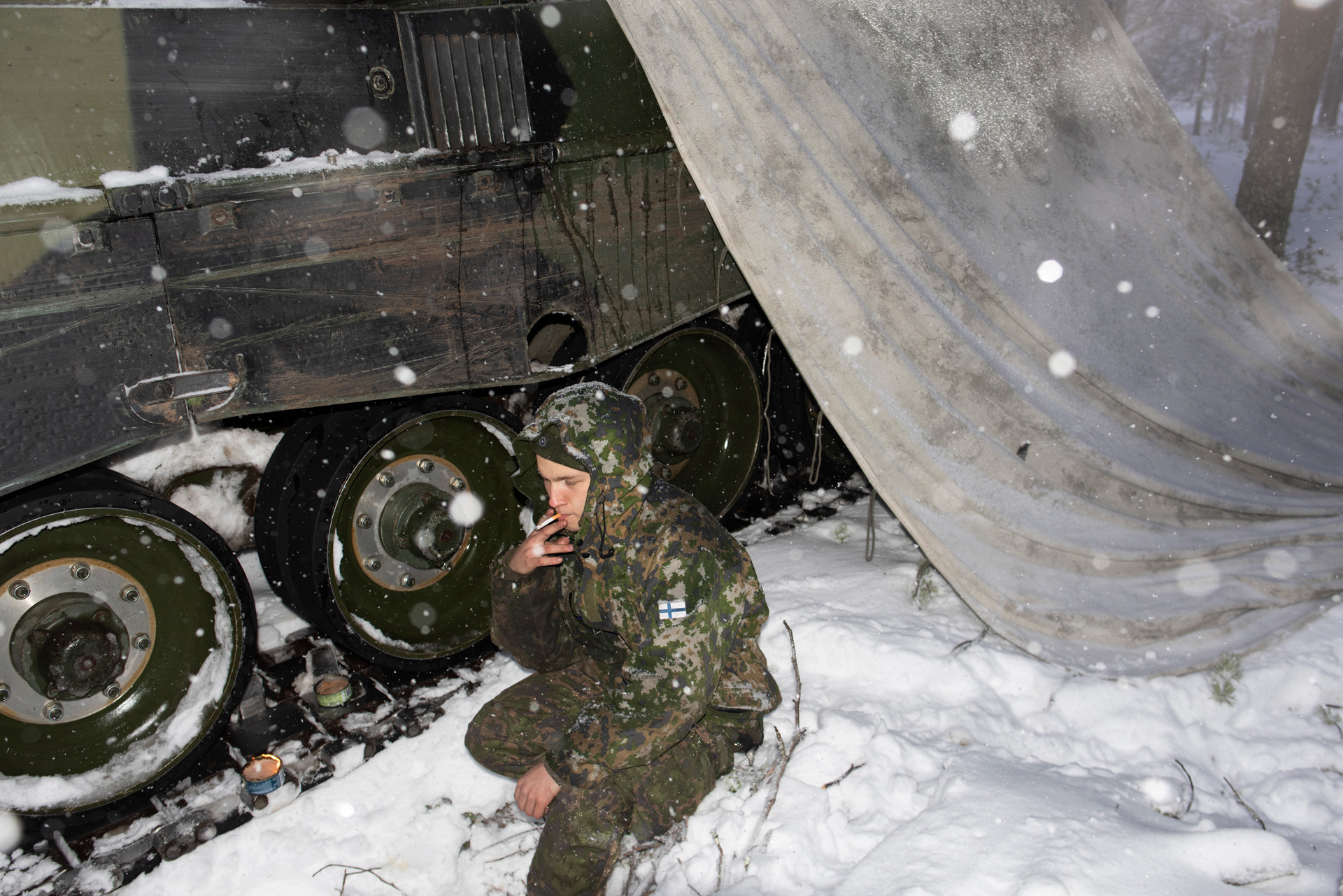A Finnish soldier wearing a green camouflage uniform crouches in the snow next to the tracked wheels of a tank that is partly covered with a white and gray tarp. He is holding a cigarette to his lips and looking down, away from the camera. Two small cans of fuel with lit flames sit on the tank track. Snowflakes fall through the air.