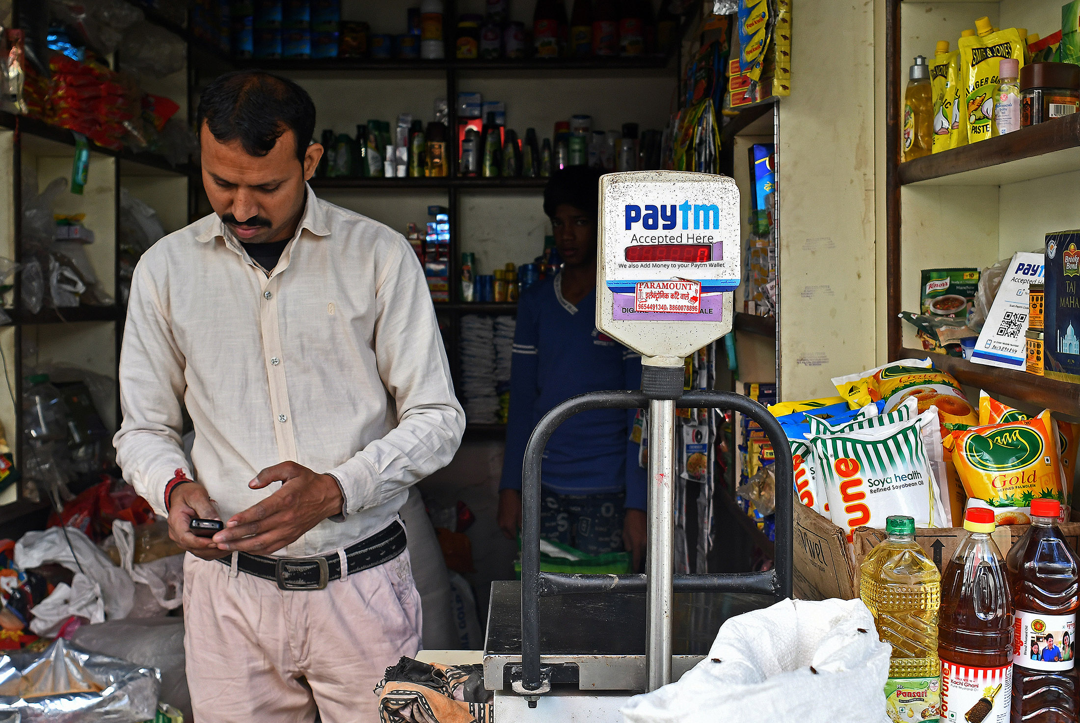 A man using a mobile device stands next to a sign for the PayTM online payment method, operated by One97 Communications Ltd., at a grocery store in Delhi, India, on Friday, Nov. 25, 2016. India's government is grappling with a furor over a rupee note ban that invalidated 86 percent of the currency in circulation.
