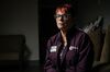 Karen Chrapek sits in a dark room. She has red hair with bangs and is wearing glasses, a deep purple button-down shirt that reads “Volusia Recovery Alliance” on the chest and her work nametag.