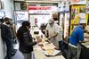 Customers line up behind the counter at a pizza restaurant. Slices of pizzas are laid across it.