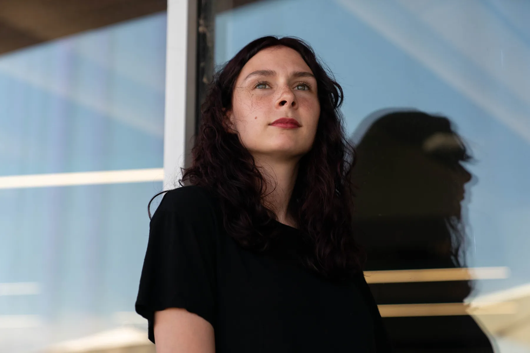 Mujer joven con cabello oscuro y rizado, vestida con una camiseta negra, mirando hacia el horizonte con una expresión serena. Está de pie frente a una ventana de vidrio, que refleja su silueta ligeramente. La escena muestra luz natural, con un cielo despejado en el fondo y líneas de luz que cruzan el cristal.