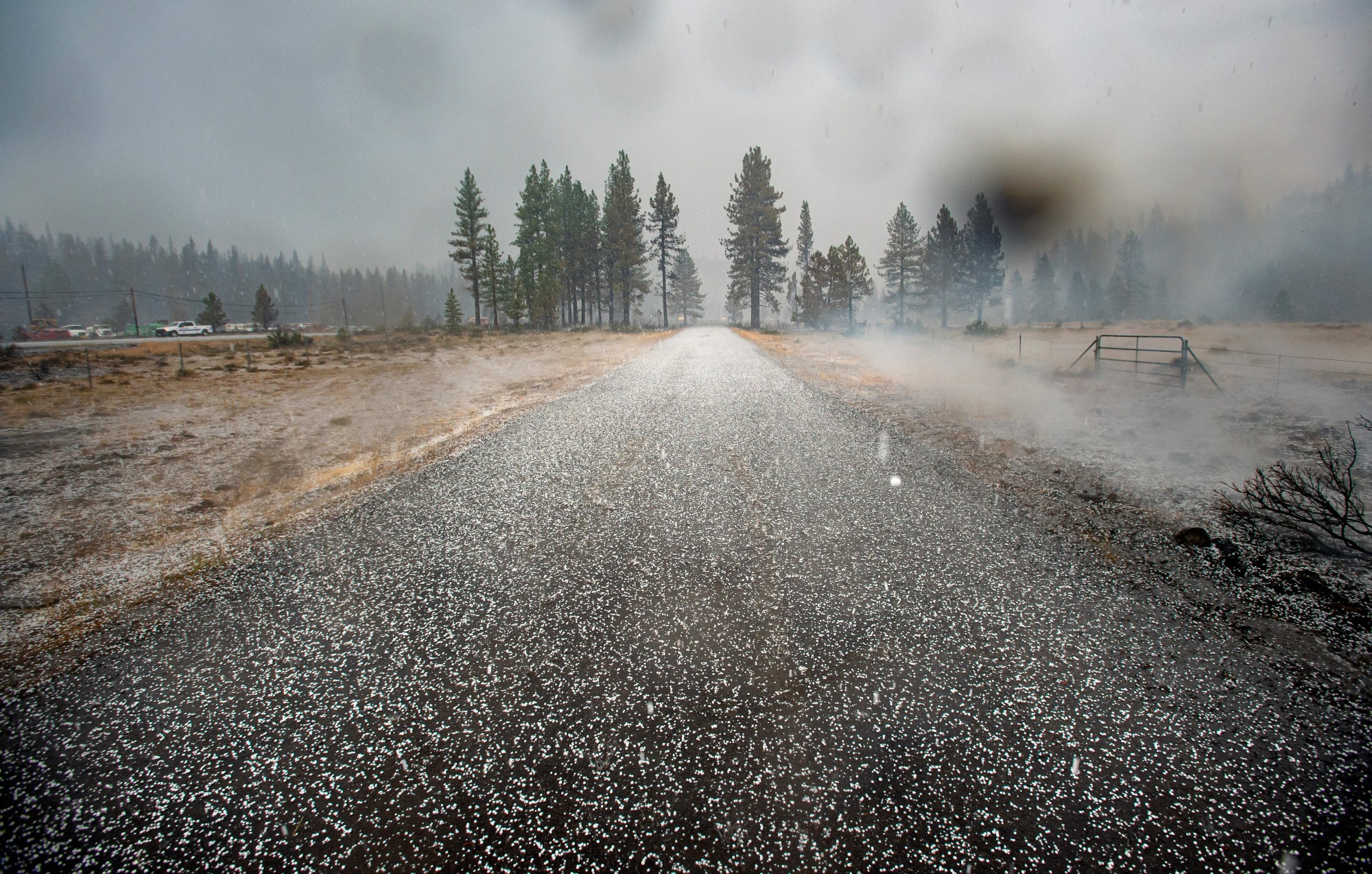 A highway covered in hail on a cloudy day. Mountains and tall trees can be seen in the distance.