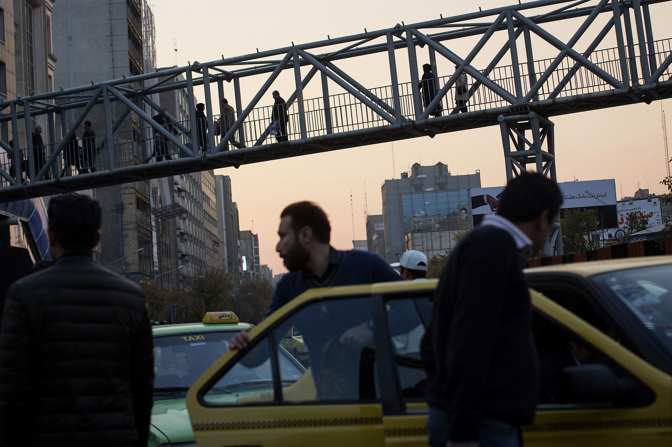 A commuter exits a taxi as pedestrians use a footbridge overhead on 7 E Tir Square in Tehran, Iran.
