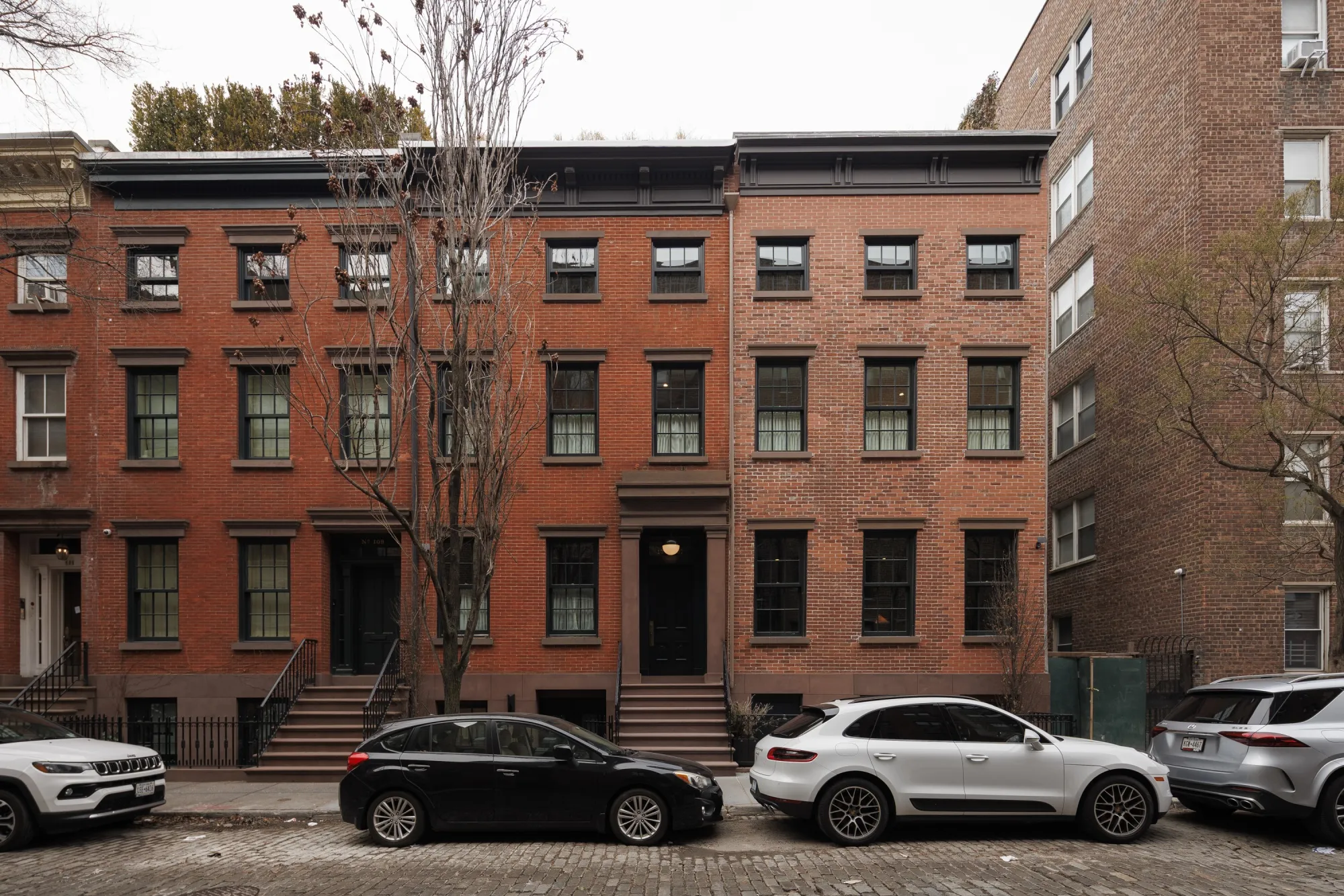 The exterior of a red-brick double-wide mega-mansion with cars parked infront of it on a cobble road.