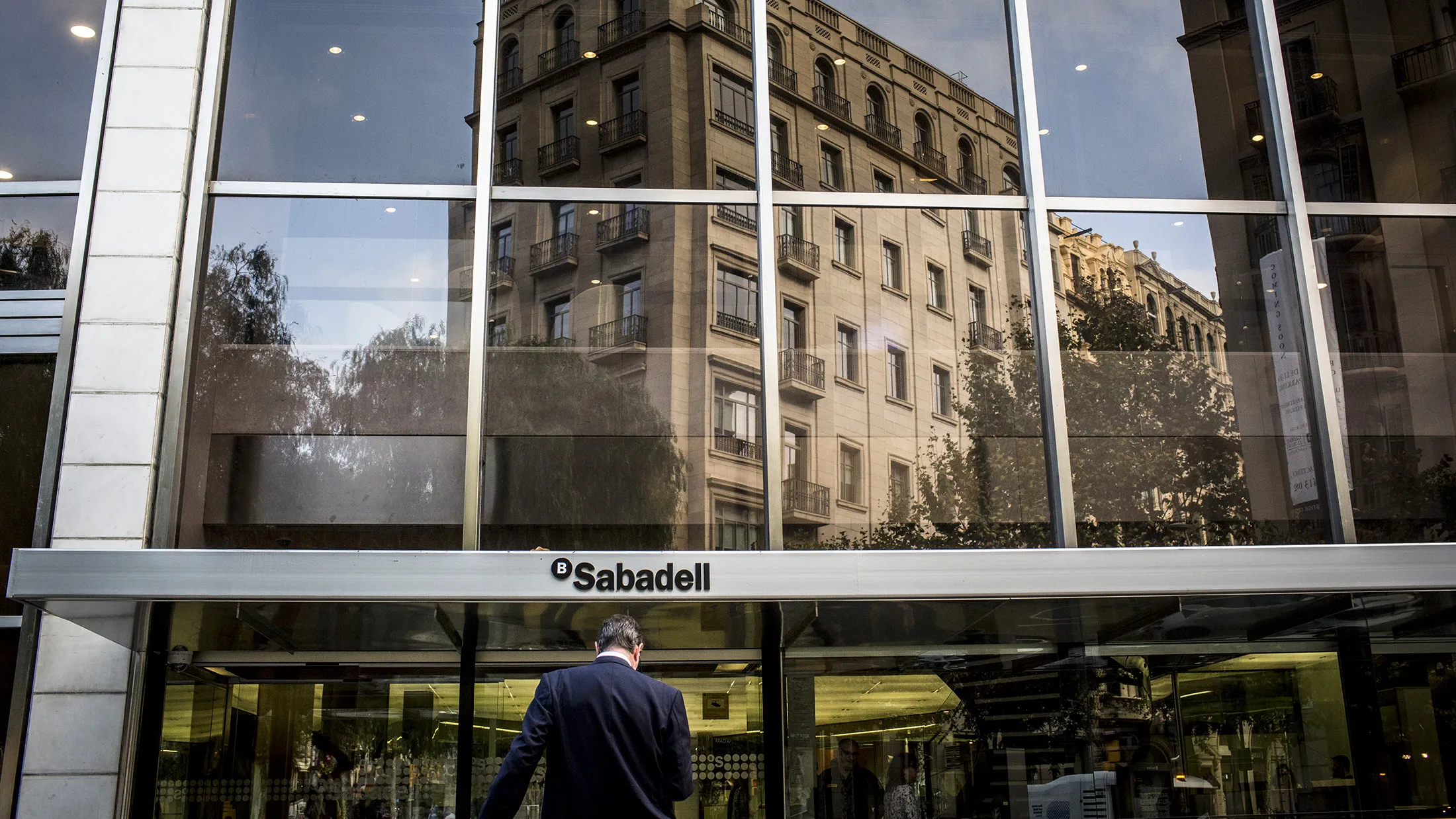 A pedestrian enters the headquarters of Banco de Sabadell SA in Barcelona, Spain, on Friday, Oct. 6, 2017. Catalan lenders such as CaixaBank SA and Banco Sabadell SA -- which have spent years seeking to reduce political risk through expansion -- have once again been pulled into the regions volatile politics following Catalonias threat of unilateral independence.
