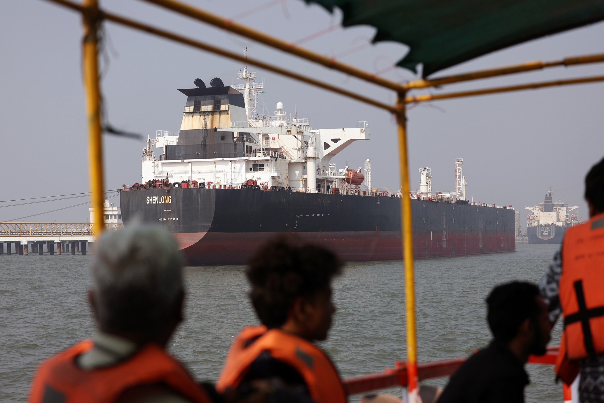 Viewed from the perspective of passengers wearing orange life jackets on a small ferry, the massive oil tanker "SHENLONG" sits docked at a harbor pier under a hazy sky.