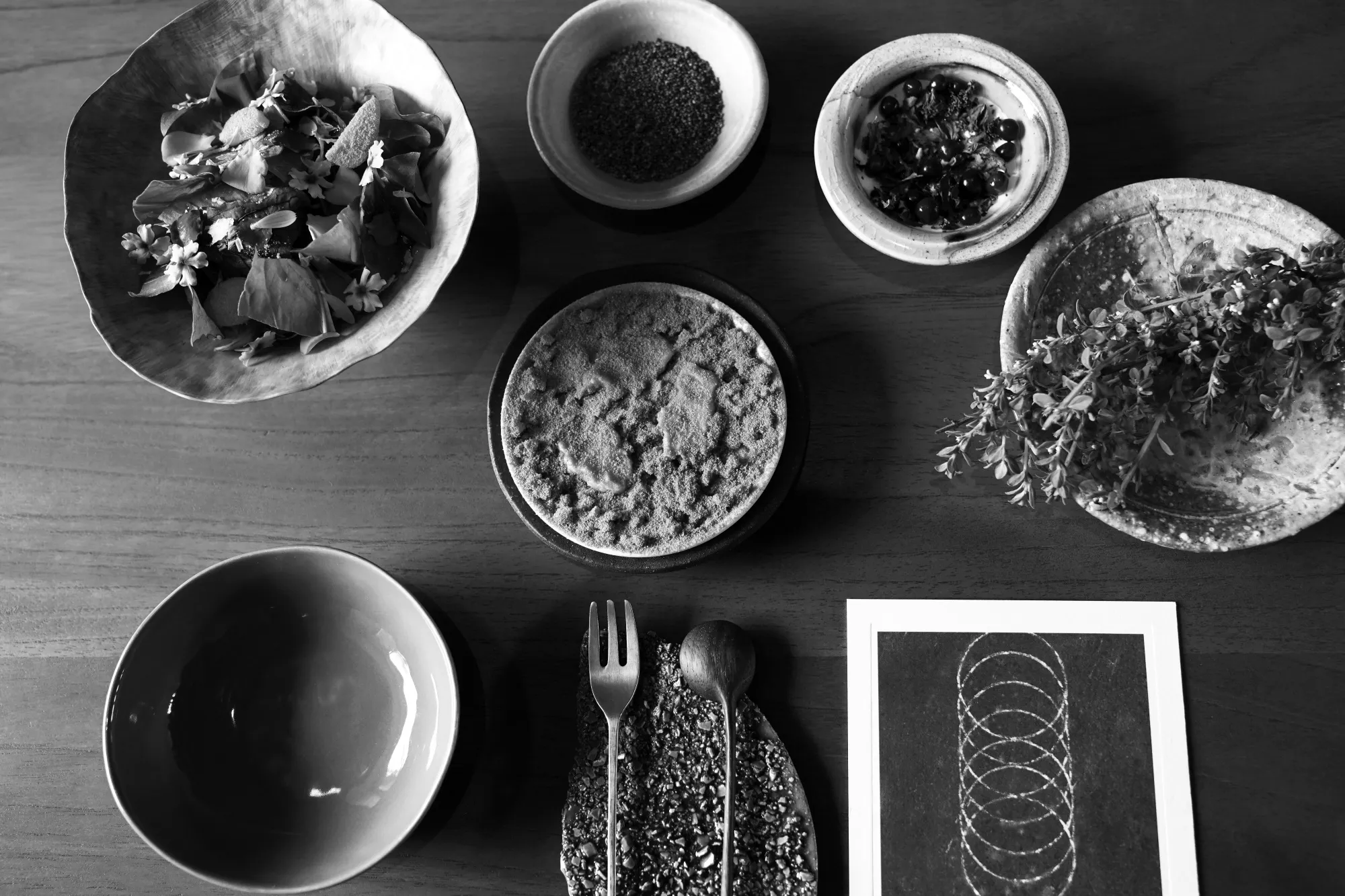 A black and white image MIL's "Highland" course, featuring lamb and cushuro all presented individual bowls with a fork and spoon resting on a textured plate in the center.