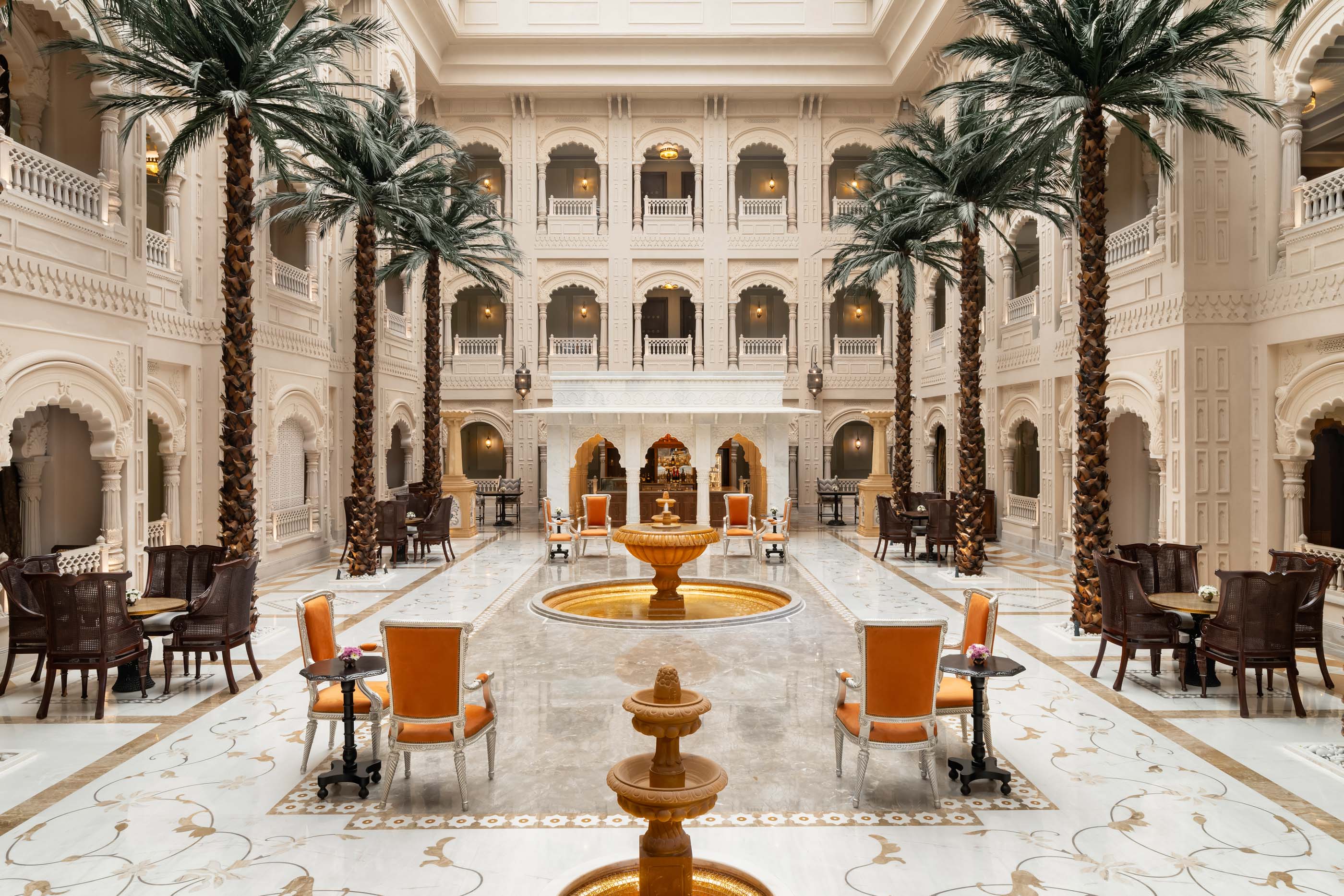 Interior of the Raffles Hotel in Jaipur with daylight illuminating the off-white central hall surrounded by three floors of decorative archway-rimmed walkways overlooking tall palm trees, lounge chairs, marble floors and golden central fountain in the middle