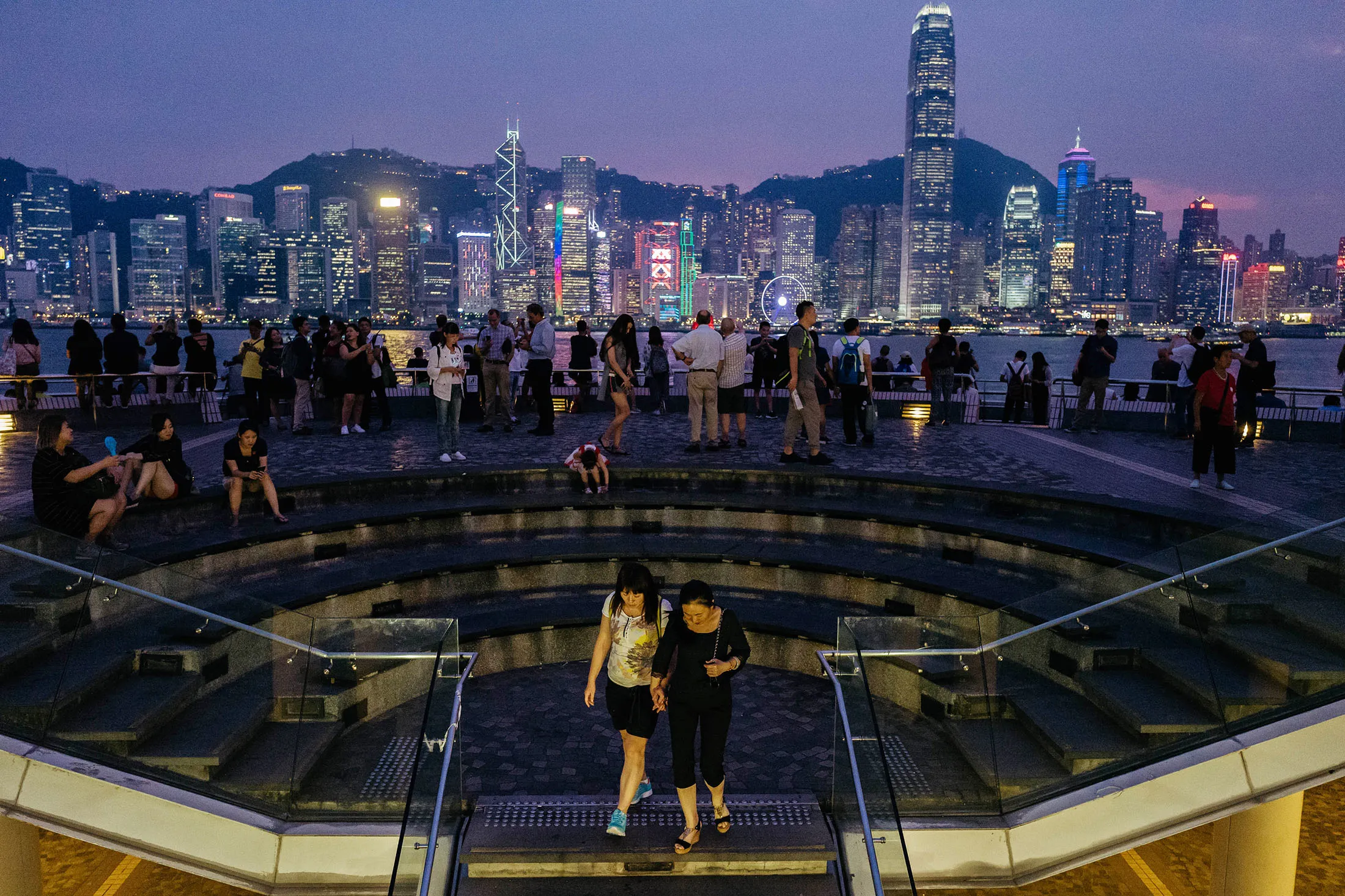 People look out from the waterfront as buildings across the Victoria Harbour stand illuminated at night in Hong Kong.
