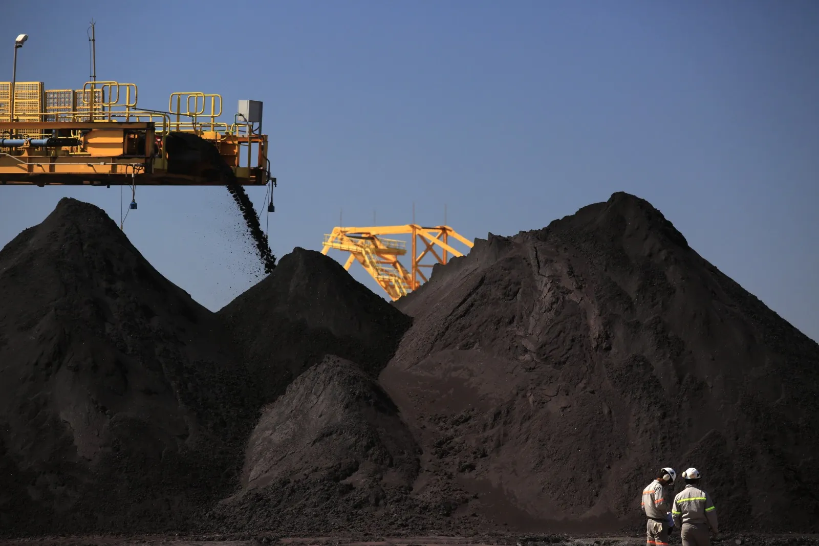 Workers stand, bottom right, as iron ore is transported and dropped into stockpiles in the Ferroport yard at the Acu Port in Sao Joao da Barra, Brazil.