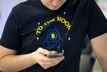 An attendee wearing a t-shirt decorated with a bitcoin rocket illustration and the words 'To the Moon' checks his smartphone at the CrytoSpace conference.
