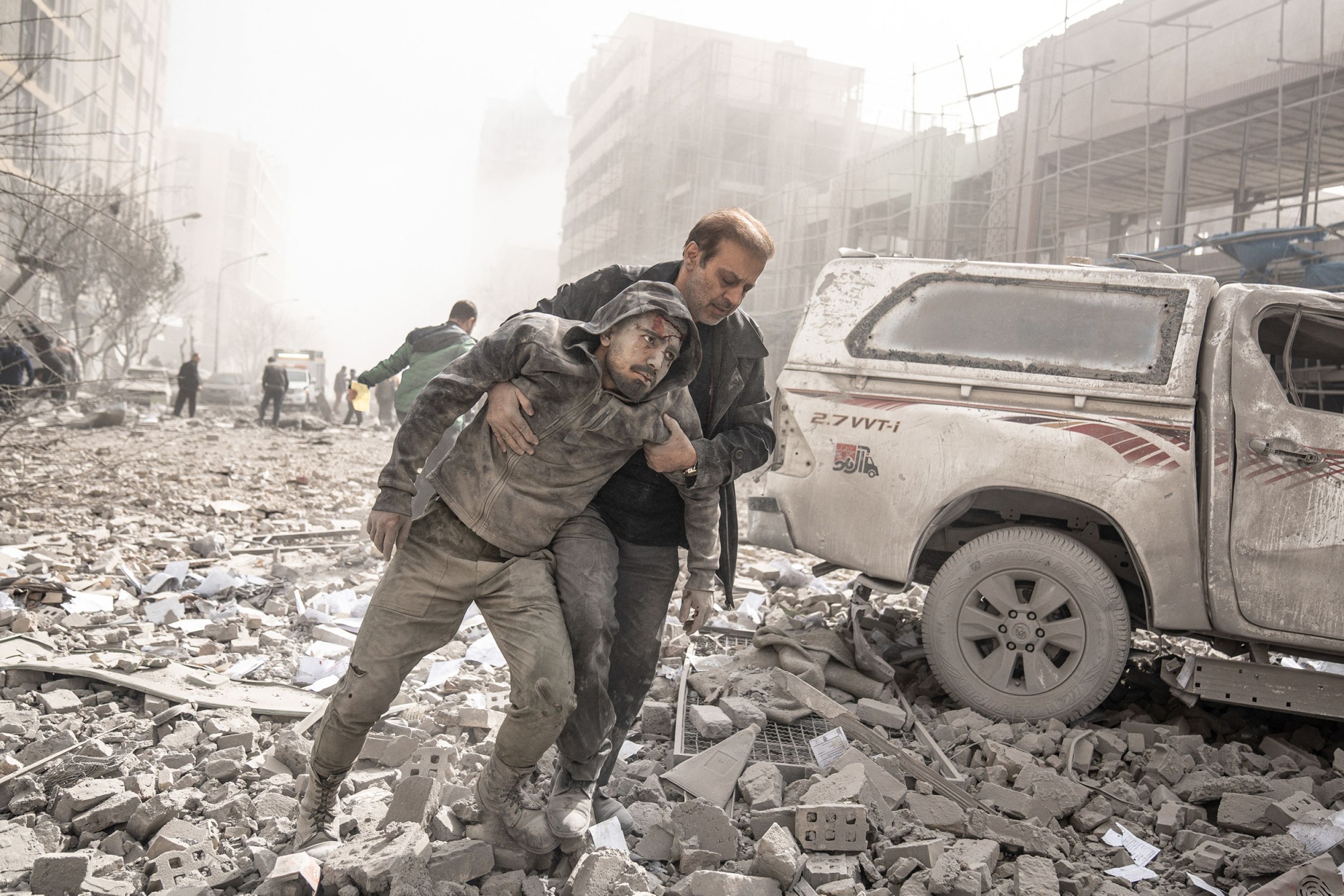 Two men covered in dust walk over rubble in a city street between destroyed buildings. One man is helping the other to walk and there is blood on his forehead.