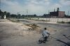 A boy wearing a gray camoflage shirt is rounding the corner on a bicycle; the dome is gone, but there is still a wire fence without concertina wire