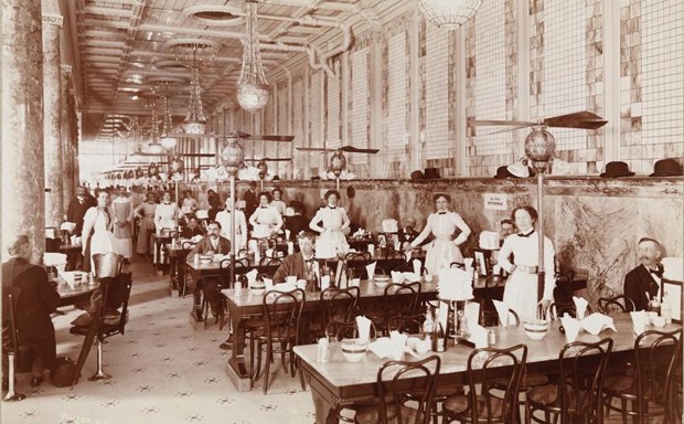 A large dining room in Childs Restaurant; long communal dining tables and wait-staff visible, circa 1899. 