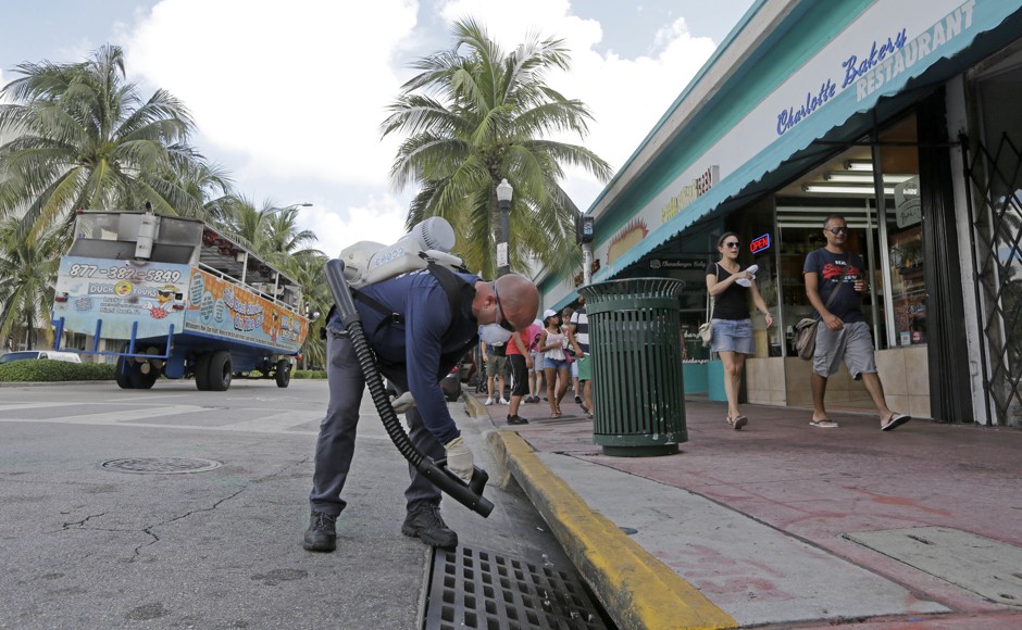A mosquito control inspector sprays a chemical mist into a storm drain as an amphibious tour bus passes by in Miami Beach.