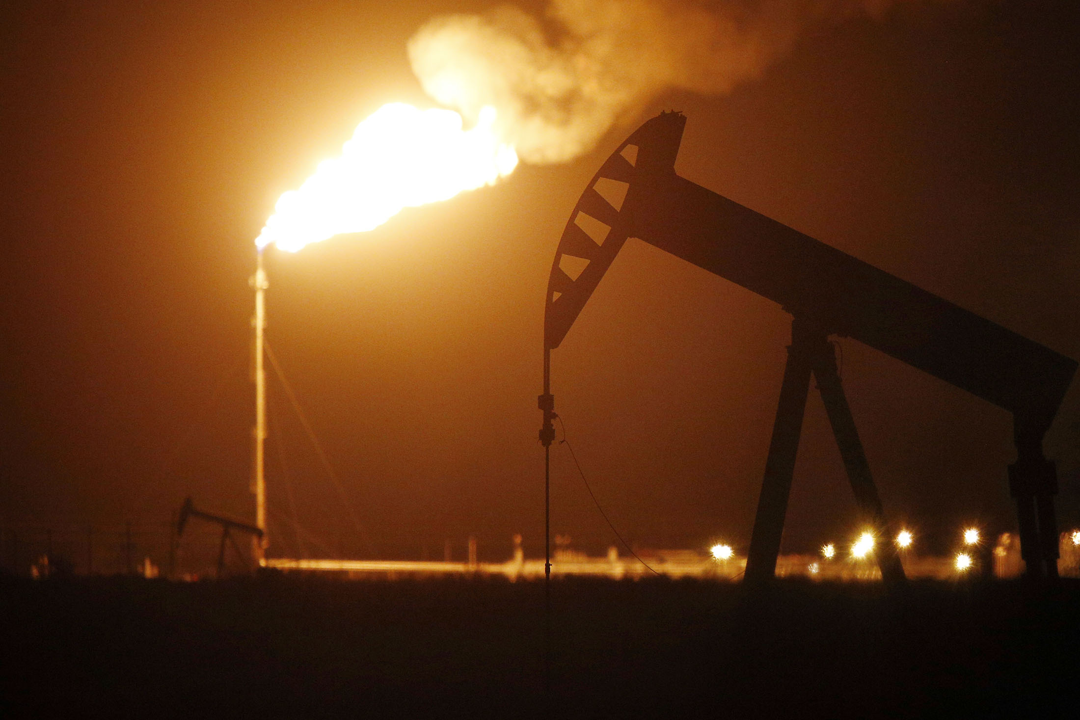 The silhouette of an electric oil pump jack is seen near a flare at night in the oil fields surrounding Midland, Texas, U.S., on Tuesday, Nov. 7, 2017. Nationwide gross oil refinery inputs will rise above 17 million barrels a day before the year ends, according to Energy Aspects, even amid a busy maintenance season and interruptions at plants in the U.S. Gulf of Mexico that were clobbered by Hurricane Harvey in the third quarter.