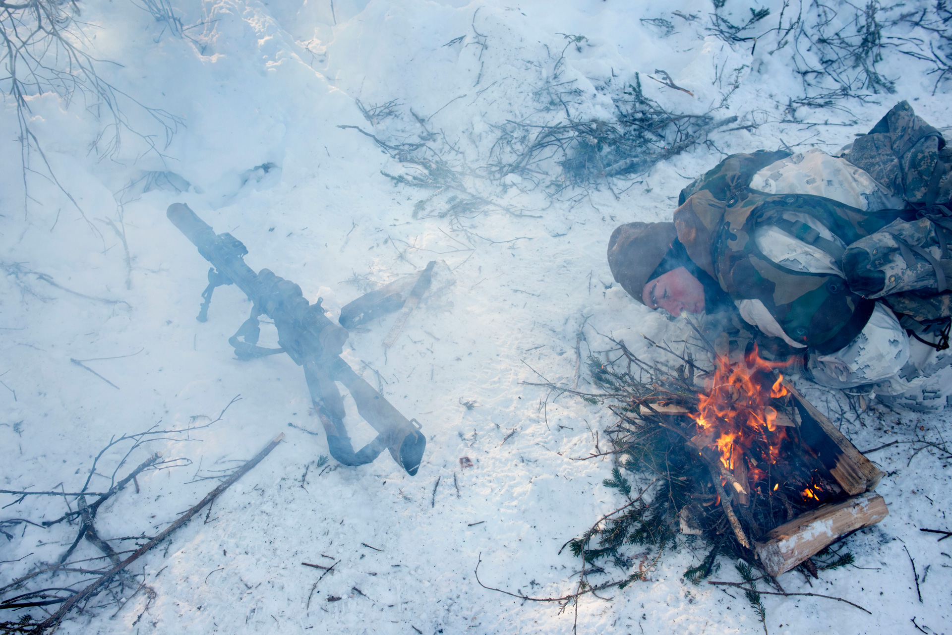 A US soldier wearing a mixture of white and khaki camouflage lies on their side on rough, snow-covered ground in Finland. They are blowing on a lit campfire of pine brush and wood, with flames and smoke rising. A tripod-mounted gun sits on the ground off to their side.