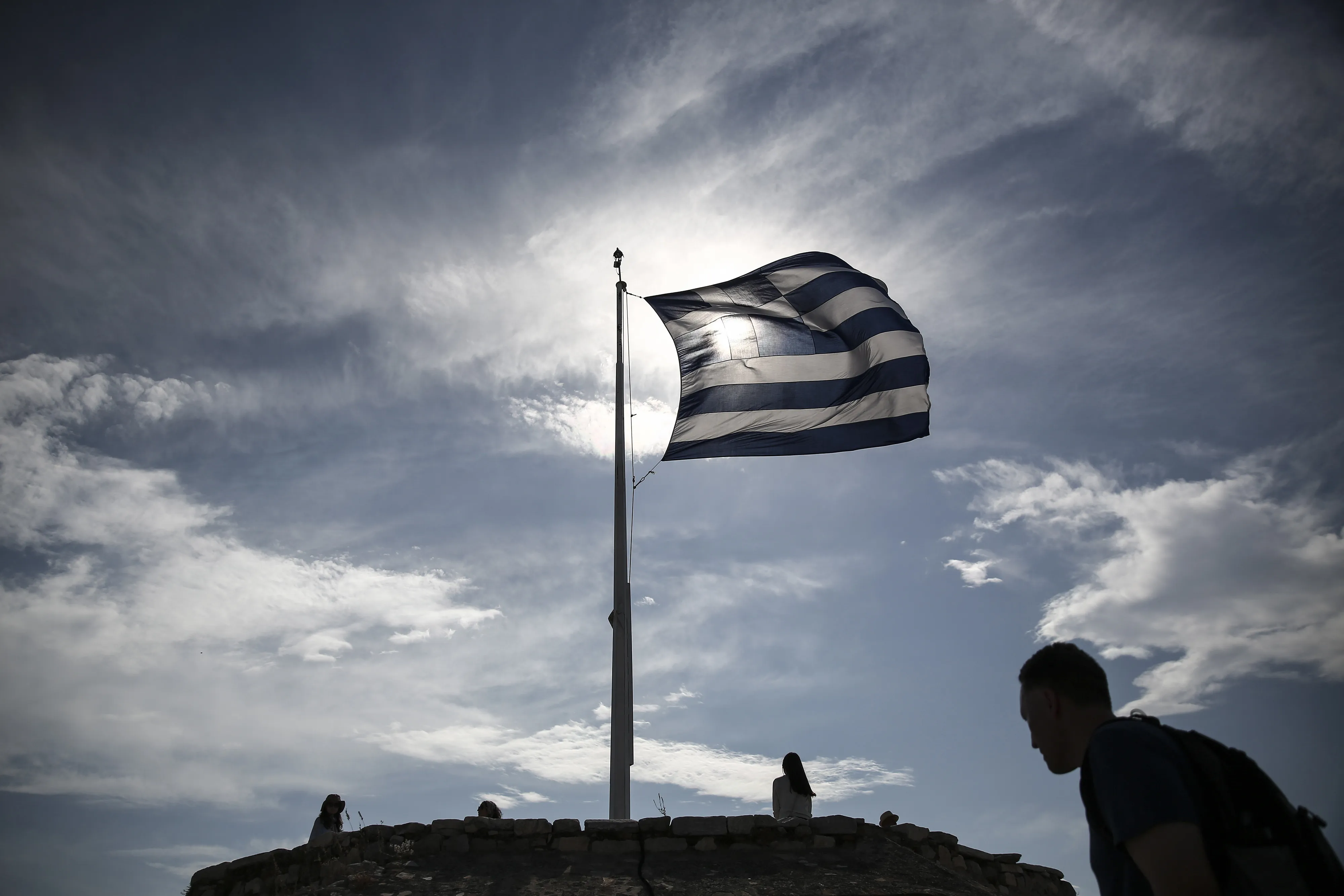 A Greek national flag flies from a flagpole above tourists visiting Acropolis hill in Athens.
