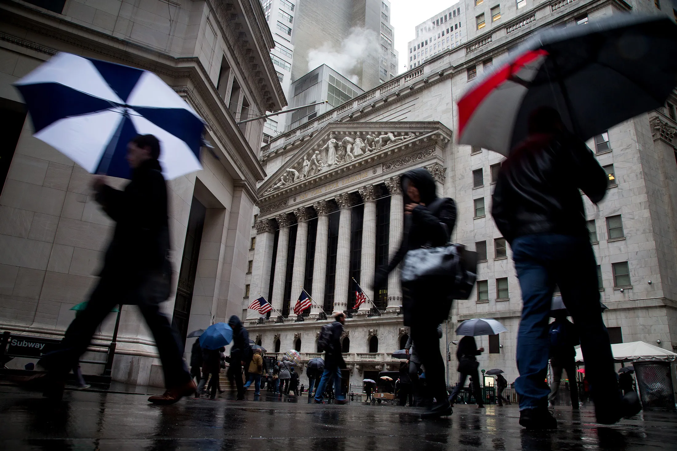 Pedestrians carry umbrellas while walking past the New York Stock Exchange (NYSE) in New York, U.S., on Monday, April 4, 2016. U.S. stocks fluctuated near their highest levels this year, with gains in health-care shares offsetting declines among consumer companies, as investors looked for fresh reasons to extend a rally.
