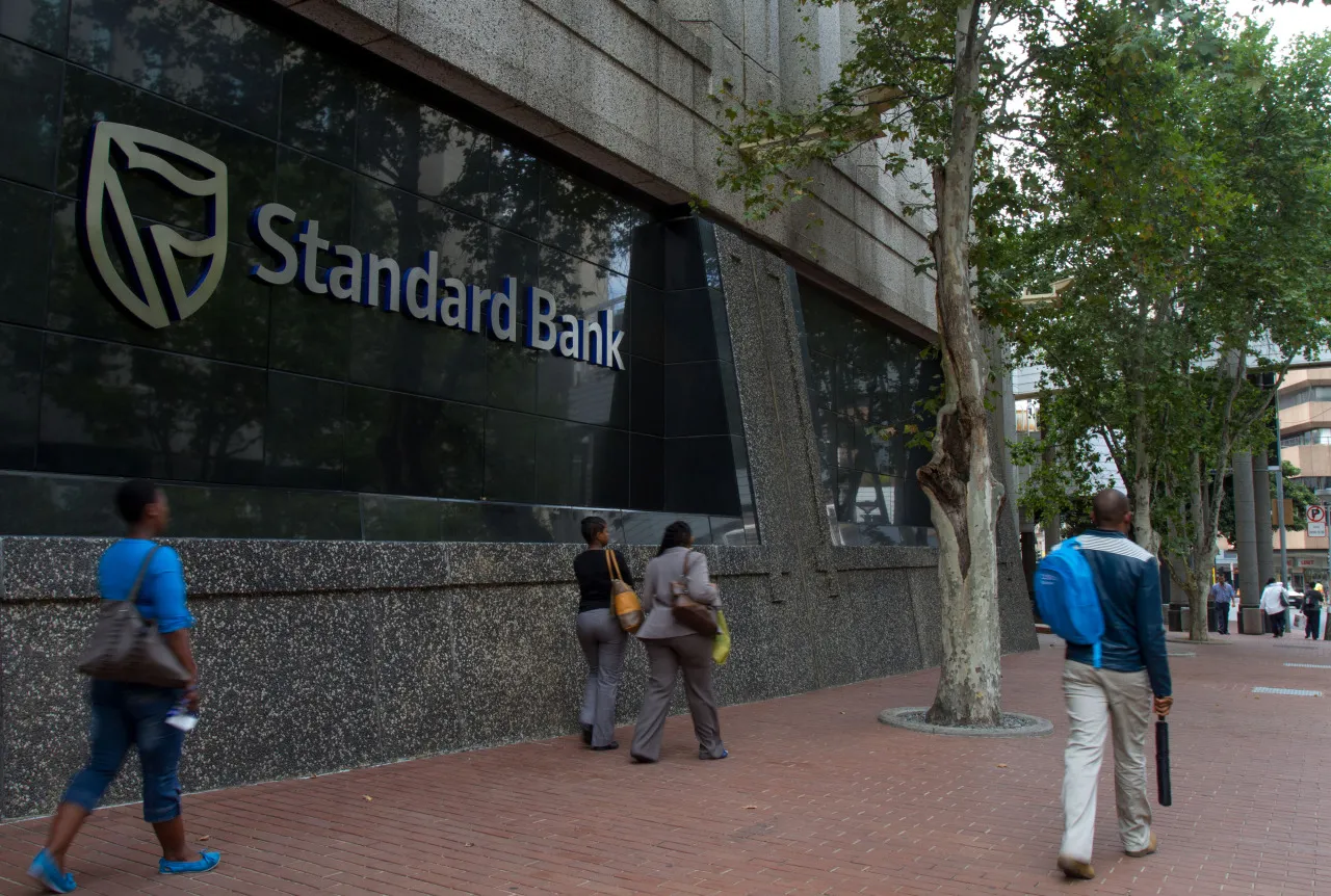 Pedestrians pass a logo outside the headquarters of Standard Bank Group Ltd. in Johannesburg, South Africa, on&nbsp;Dec. 19, 2013.