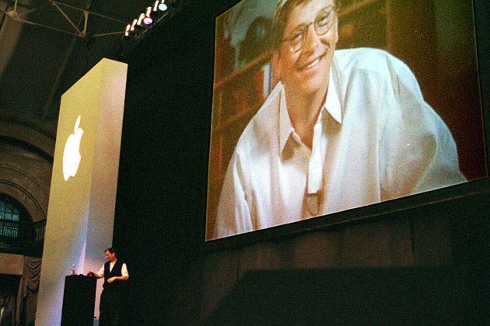 Steve Jobs stands at a podium as Bill Gates appears on a video screen as he addresses the MacWorld convention praising the new alliance between Apple and Microsoft on Aug. 6, 1997 in Boston