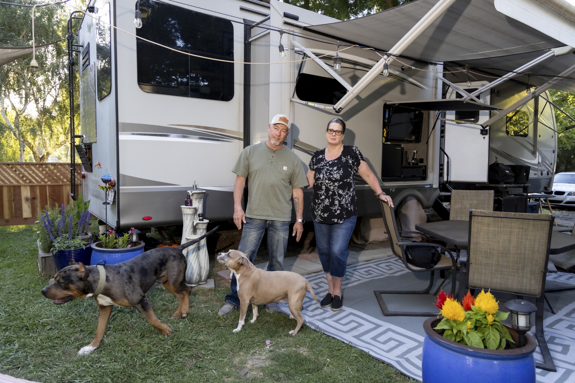 Husband and wife hold hands surrounded by their three dogs, RVs and a brown fence line the background.