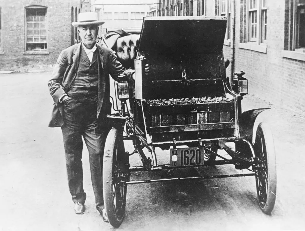 Thomas Edison with his first electric car, the Edison Baker. He’s holding one of the batteries used to power the vehicle.&nbsp;