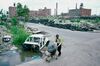 Man passing the metal skeleton of a car, pushing a shopping cart filled with garbage bags, on a street lined with mountains of tires and hollowed-out factories