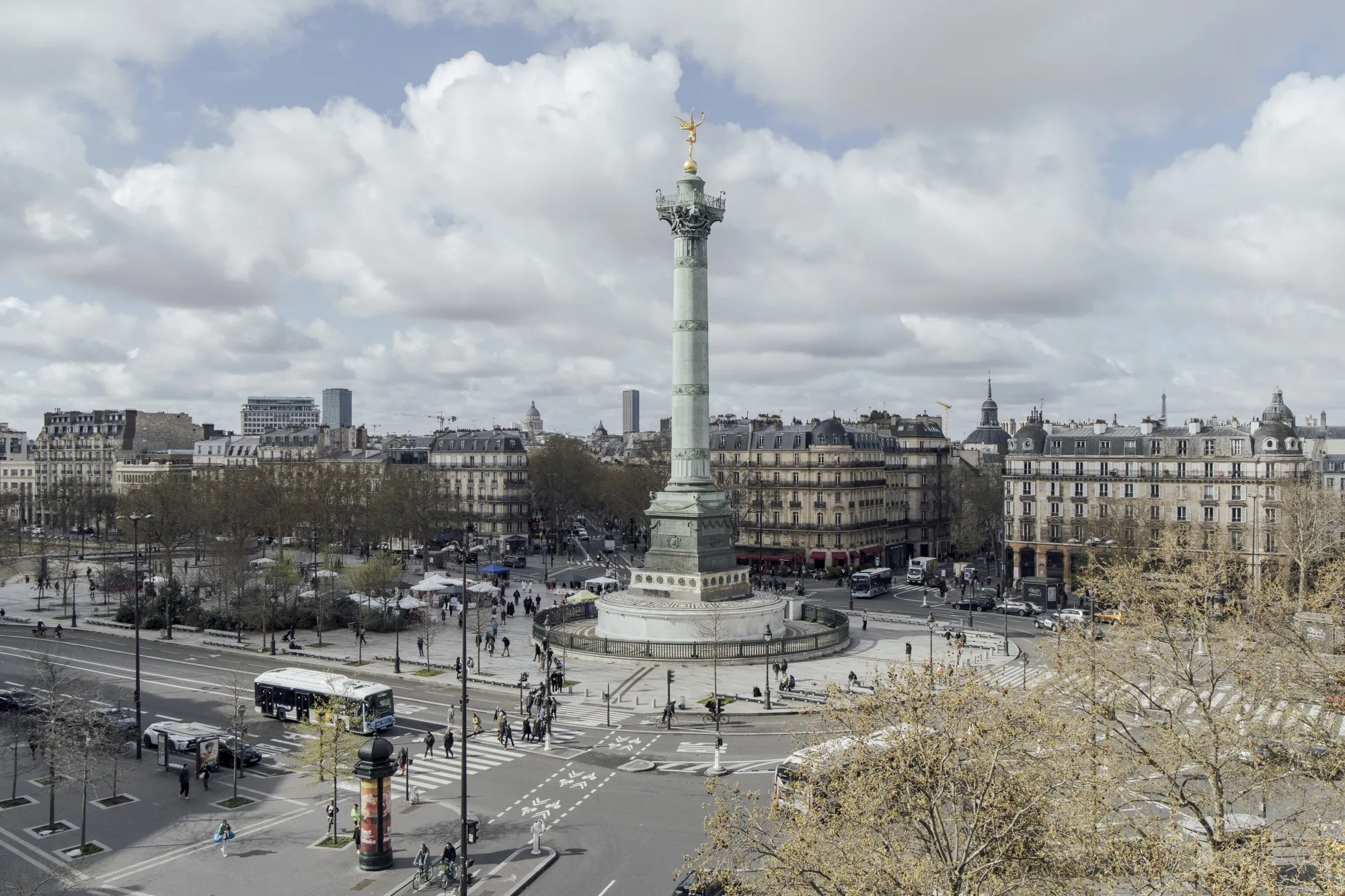 An elevated wide shot of the Place de la Bastille in Paris on a bright but cloudy day in March. In the center stands the Colonne de Juillet, a tall pillar topped with a golden winged statue. At the base of the column, people walk through a paved plaza and an open-air market with white tents. The foreground shows a wide city street with a bus, a crosswalk, and several pedestrians. In the background, traditional Parisian stone apartment buildings line the streets under a sky filled with white clouds.