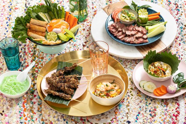Table of food including skewers, rice with shrimp and drinking glass, all placed in gold serving tray. Fried roll with assorted vegetables in seafoam green bowl, an d other meats an vegetables on serving trays.