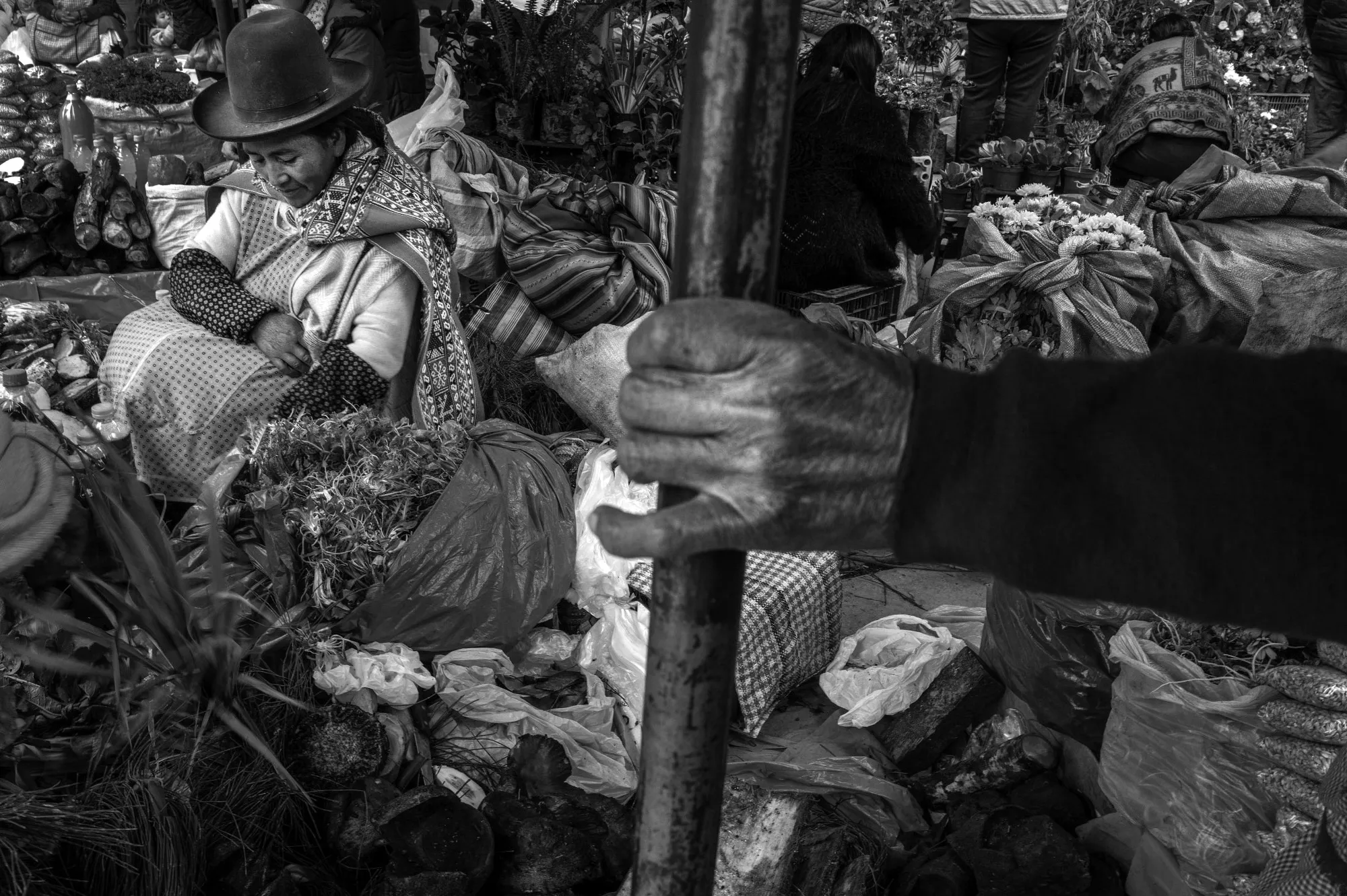 A black and white image of Hilda Tejada, left, at the Urubamba farmers market. A hand holding a post is seen in the foreground.