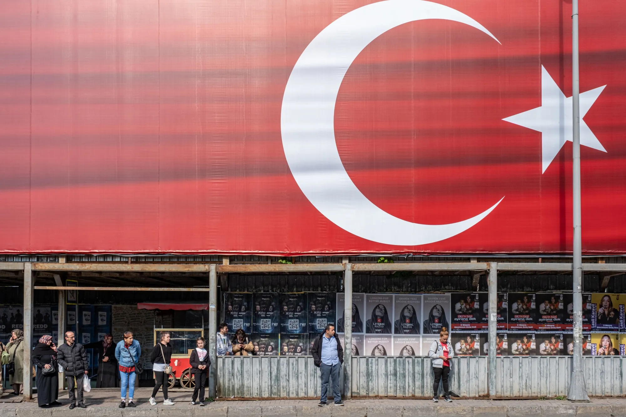 Residents wait at a bus stop under a large Turkish flag building hoarding in Istanbul.